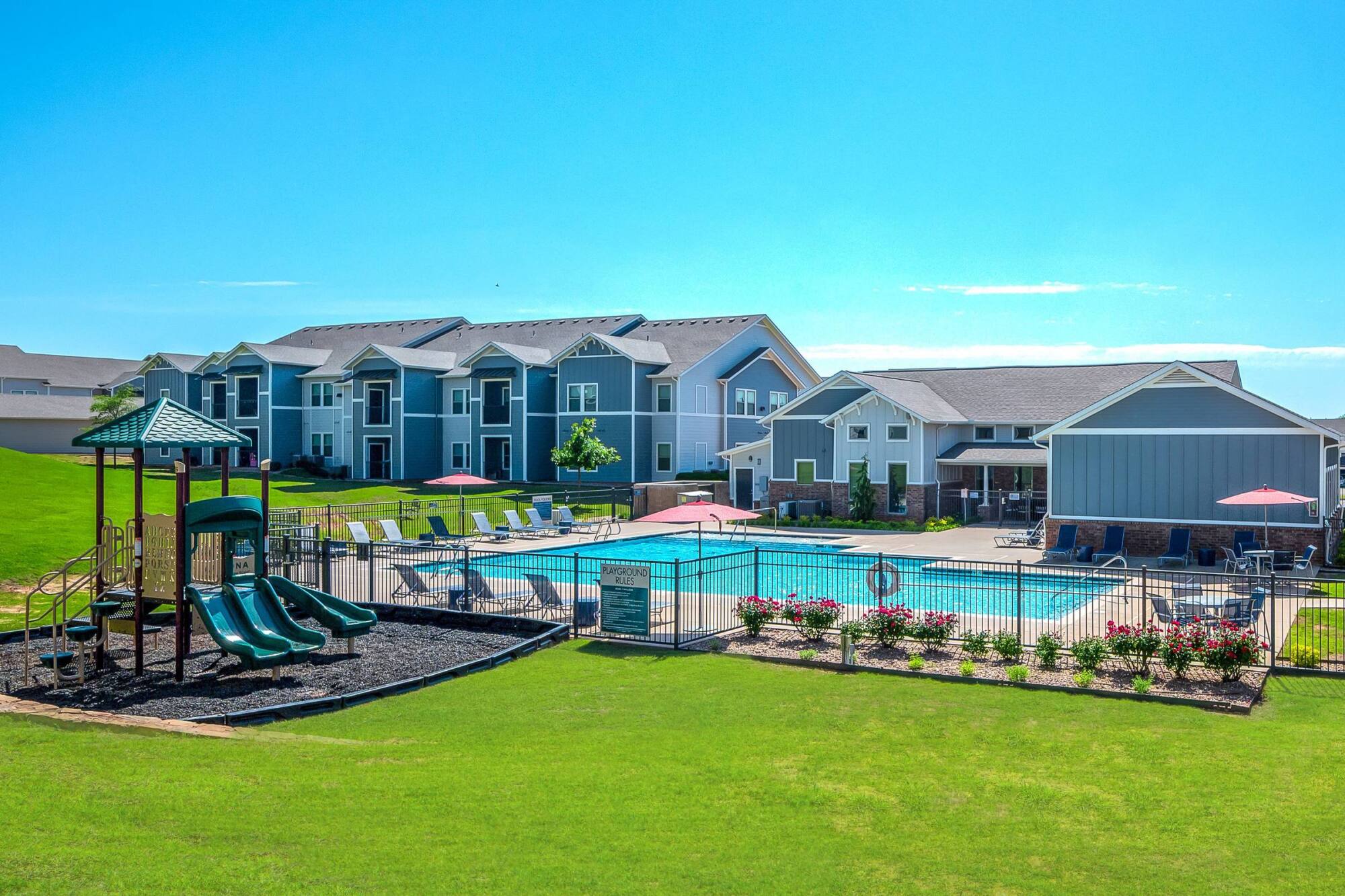 Apartment complex with playground, green lawn, lounge chairs, and a fenced outdoor pool under a clear blue sky.