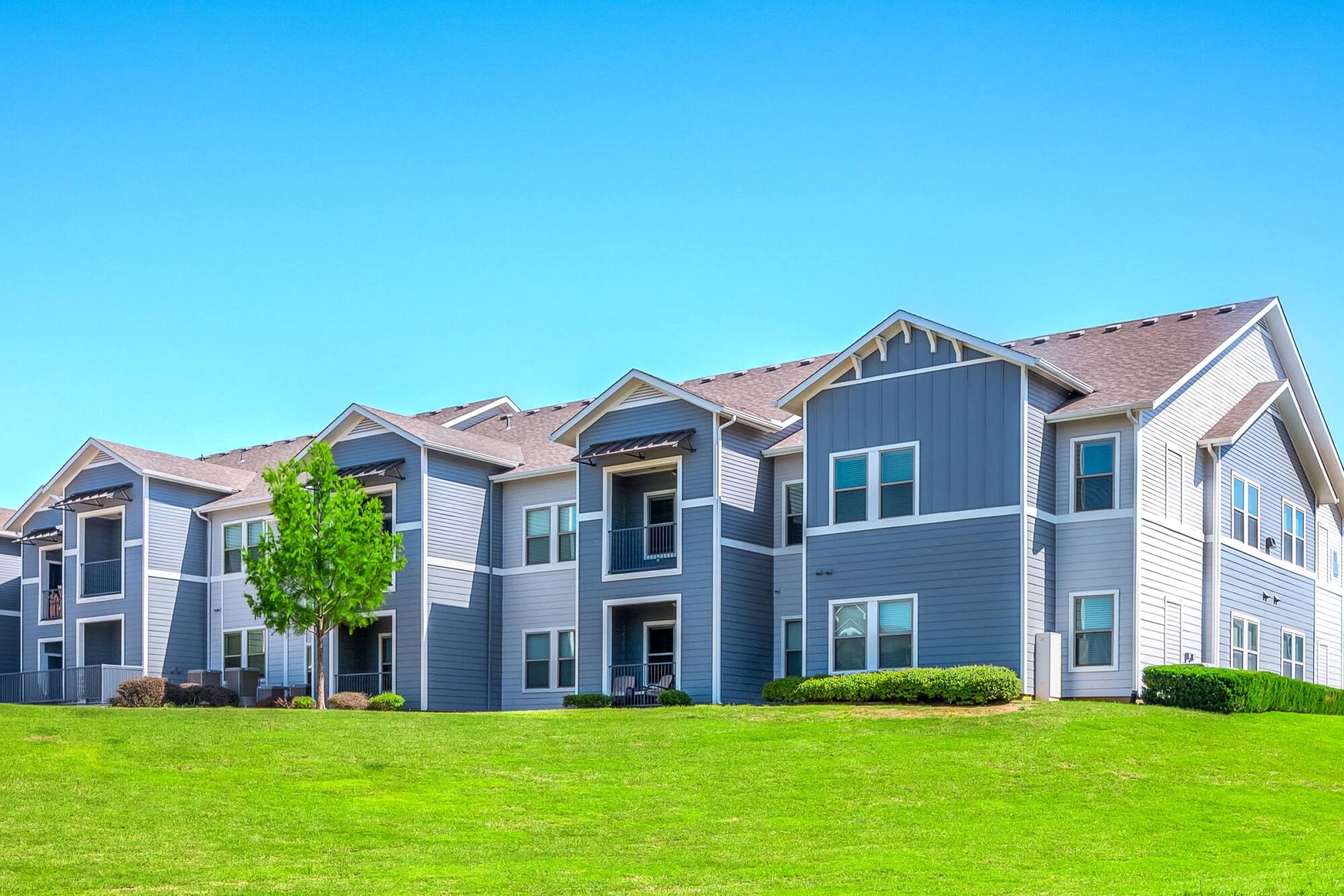 Modern gray and white apartment building with balconies, surrounded by green grass and a clear blue sky.