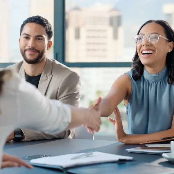 Smiling woman in glasses shakes hands with another person across a table during a meeting.